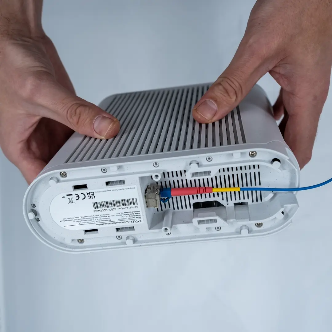 En person holder ruteren opp-ned og viser hvordan SFP-modulen, med den tilkoblede blå fiberkabelen, settes inn i kontakten på undersiden av enheten.